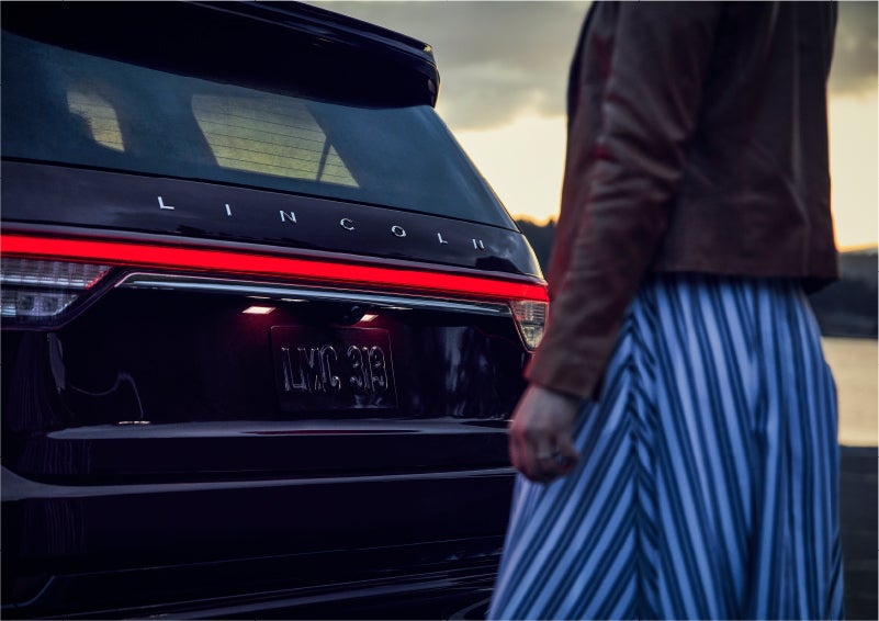 A person is shown near the rear of a 2023 Lincoln Aviator® SUV as the Lincoln Embrace illuminates the rear lights | Apple Lincoln Apple Valley in Apple Valley MN
