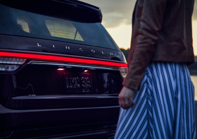 A person is shown near the rear of a 2024 Lincoln Aviator® SUV as the Lincoln Embrace illuminates the rear lights | Apple Lincoln Apple Valley in Apple Valley MN